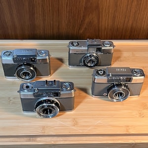 May include: Four vintage Olympus-Pen cameras are displayed on a wooden surface. The cameras have a silver and grey colour scheme with black lenses. The text "Olympus-Pen" is visible on some of the cameras.