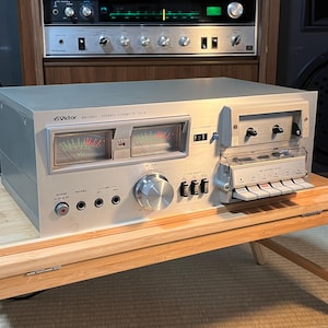 Könnte beinhalten: Silbernes Victor KD-A50 Stereo-Kassettendeck mit VU-Metern und einem Kassettenfach. Das Deck hat verschiedene Knöpfe und Regler. Das Deck steht auf einem Holztisch.