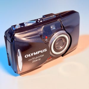 Puede incluir: Una cámara negra Olympus Stylus Epic Zoom 80 con tapa de lente deslizante, flash integrado y un objetivo con rango de zoom de 38-80 mm. El cuerpo de la cámara muestra la marca Olympus y el modelo Stylus Epic Zoom 80 en texto de color blanco.