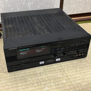 May include: A black Kenwood compact disc stereo system. The front panel displays "AUX" and various control buttons. The top has a vented design. The unit is on a light-colored woven surface.