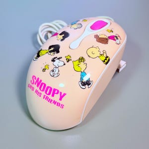 Op de afbeelding: Een licht perzikkleurige computermuis met een grafische print van Snoopy en zijn vrienden. De tekst SNOOPY AND HIS FRIENDS staat in felroze op de voorkant gedrukt. Het bevat een wit scrollwiel met een roze midden en een wit snoer.