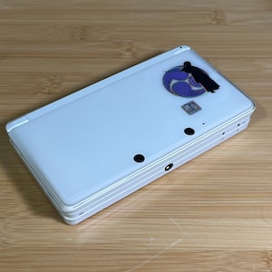 Può includere: Console di gioco portatile bianca con un adesivo decorativo con una silhouette nera e un design blu e viola. La console ha pulsanti neri e una forma rettangolare.