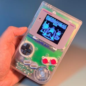 Op de afbeelding: Een doorzichtige Nintendo Game Boy handheld console met een groene interne printplaat, een zwarte D-pad, twee felrode A- en B-knoppen en twee grijze Select- en Start-knoppen. Het scherm toont de titel Super Donkey Kong GB.