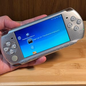 Könnte beinhalten: Eine silberne tragbare Spielkonsole mit einem Breitbildschirm, der die PlayStation Network-Oberfläche anzeigt. Das Gerät verfügt über ein Steuerkreuz auf der linken Seite, vier Aktionstasten rechts und eine Reihe von Steuertasten am unteren Rand.
