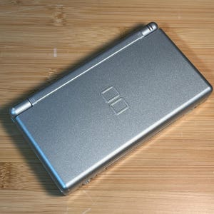 May include: A silver handheld gaming console with a clamshell design. The top cover features a metallic finish and a centered embossed DS logo. The device is rectangular with rounded edges and a hinge running along the top side of the unit.