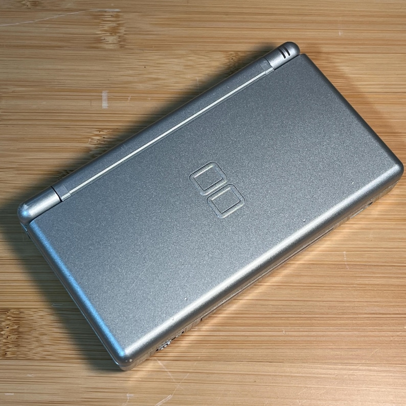 May include: A silver handheld gaming console with a clamshell design. The top cover features a metallic finish and a centered embossed DS logo. The device is rectangular with rounded edges and a hinge running along the top side of the unit.