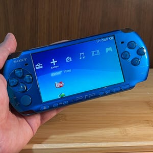 以下が含まれることがあります： 青いソニーPSPポータブルゲーム機。デバイスには、アイコン付きのメニューを表示する画面があります。コンソールには、Dパッド、ボタン、ジョイスティックがあります。画面には時刻が1/1 0:00と表示されます。