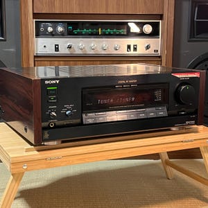 May include: A vintage Sony Digital AV Master receiver with a dark wood finish. The faceplate is black with a display that reads "TUNER". It has various buttons and knobs, including a large volume control. The receiver sits on a small wooden table.