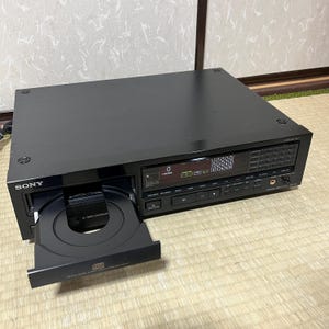 Puede incluir: Reproductor de CD Sony negro con una bandeja de disco abierta. El reproductor tiene una pantalla digital y botones de control. La parte frontal del reproductor tiene el logotipo de Sony. La bandeja de disco está abierta, revelando el compartimento del CD. El reproductor está sobre una superficie clara.