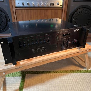May include: Black APA multi-compact amp model MR-400. The front panel features various knobs, switches, and level indicators. The amp is on a small wooden table. The background includes a wooden cabinet and speakers.