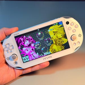 Könnte beinhalten: Eine weiße Sony PS Vita Handheld-Spielkonsole mit einem hellen Bildschirm, der eine farbenfrohe Anime-Oberfläche mit Charakteren in Pink und Gelb anzeigt. Das Gerät verfügt über ein Steuerkreuz, Analogsticks und Standard-Aktionstasten.