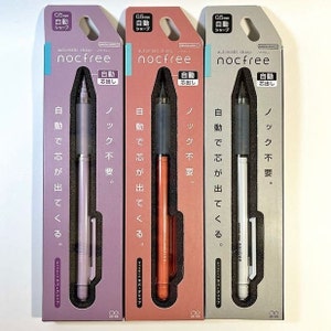 Könnte beinhalten: Drei automatische Bleistifte in Einzelverpackungen. Die Stifte sind silber, orange und weiß. Die Verpackung ist grau mit japanischem Text, der "0.5mm automatic sharp nocfree" lautet.