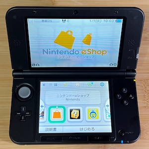 Può includere: Console di gioco portatile Nintendo 3DS nera, aperta per visualizzare l'interfaccia Nintendo eShop. Lo schermo mostra il logo eShop e testo giapponese. Lo schermo inferiore mostra icone di app e altro testo giapponese.