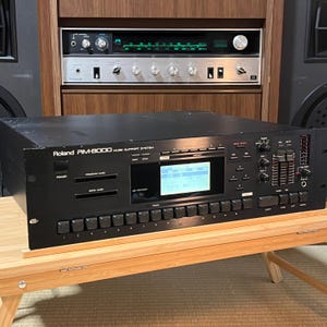 Peut inclure: Un système de support musical Roland RM-8000 noir, avec un écran d'affichage et de nombreux boutons et molettes. L'appareil est posé sur une table en bois, avec un récepteur stéréo en arrière-plan.