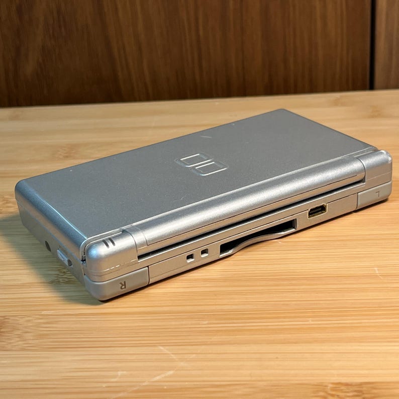 May include: A silver handheld video game console with a clamshell design. The top cover features an embossed DS logo. The side profile shows an R shoulder button, a game cartridge slot, a charging port, and a small speaker grille opening.