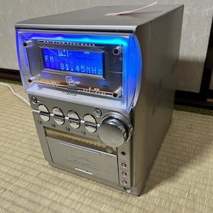 Puede incluir: Sistema estéreo compacto de color plateado con una pantalla iluminada en azul que muestra "FM 81.45MHz". El estéreo tiene varios botones y una bandeja de CD. La marca "Victor" es visible.