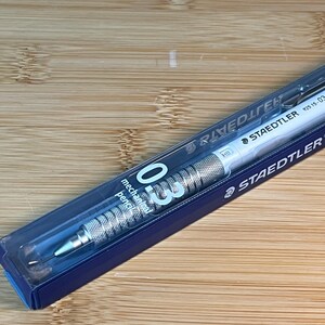 Peut inclure: Un crayon mécanique Staedtler 925 15-03 dans son emballage d'origine. Le crayon a un corps blanc et une prise en main argentée et texturée. L'emballage est un étui en plastique transparent avec un manchon en carton bleu. Le crayon est étiqueté "0.3 mechanical pencil".