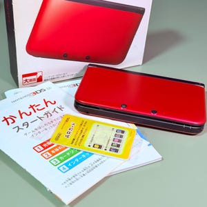 Puede incluir: Una consola de juegos portátil Nintendo 3DS LL roja y negra con su caja original, una guía de inicio rápido impresa y una tarjeta AR amarilla. La consola está cerrada, mostrando una cubierta superior roja brillante y una base negra.