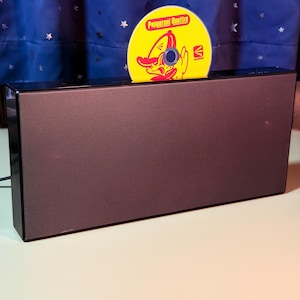 Peut inclure: Une enceinte rectangulaire noire avec une façade en maille. Un disque compact jaune est inséré dans la fente supérieure, présentant un graphique rouge d'un personnage de dessin animé et le texte Pepperoni Quattro en police rouge.