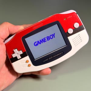 May include: A handheld Game Boy Advance video game console featuring a red, white, and black colour scheme. The screen displays the text GAME BOY in blue. The device includes a white directional pad, two white A and B buttons, and two orange buttons.