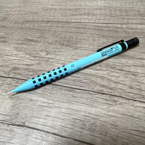 Może przedstawiać: Ołówek mechaniczny w jasnoniebieskim kolorze z czarnym wzorem siatki i napisem "SMASH 0.5 Pentel 01005 Japan" na korpusie. Ołówek leży na drewnianej powierzchni.