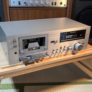 May include: A silver Technics M330 stereo cassette deck with a cassette tape inserted. The deck has various buttons, knobs, and a fluorescent meter. The deck is on a wooden surface.