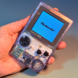 以下が含まれることがあります： 透明な紫色のゲームボーイポケット携帯ゲーム機。明るい青色の画面にはNintendoのロゴが表示されています。黒い十字キー、Aボタン、Bボタン、セレクトボタン、スタートボタンを備えており、内部の回路基板が透けて見えています。