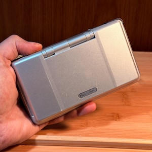 May include: A silver handheld gaming console with a rectangular, clamshell design. The exterior features a smooth, metallic finish with a prominent hinge along the top edge and the Nintendo logo embossed in the lower right corner of the lid.
