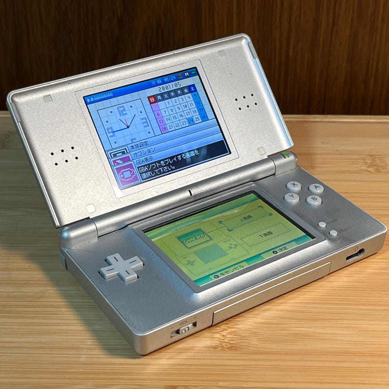 May include: A silver handheld gaming console with dual screens. The top screen displays a calendar and clock interface with Japanese text. The bottom screen shows a menu with a Game Boy icon. The device features a directional pad and four buttons.