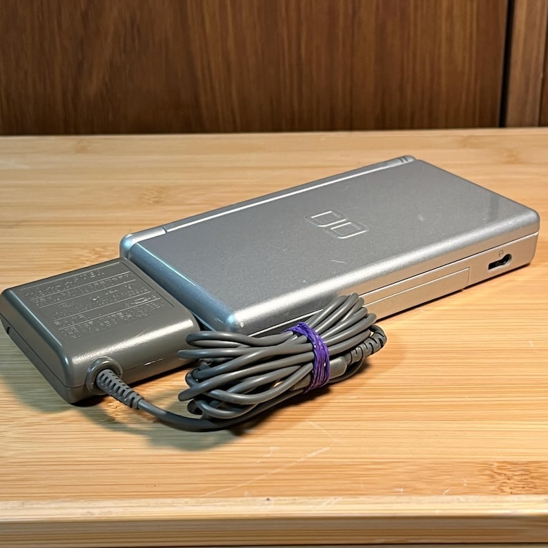 May include: A silver handheld gaming console with a closed clamshell design and the Nintendo DS logo on the lid. It is connected to a grey power adapter with a coiled cord secured by a small purple elastic band, showing a charging setup.