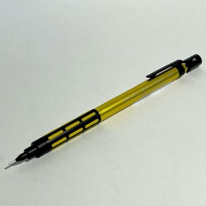 Puede incluir: Un lápiz mecánico dorado con detalles negros y un clip negro. El lápiz tiene un agarre de goma con detalles negros y amarillos. El lápiz está etiquetado como "GRAPH1000 LIMITED 0.5".