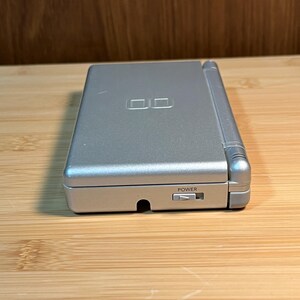 May include: A silver handheld gaming console in a closed position. The top cover features an embossed DS logo. The side edge displays a sliding power switch labeled with the word POWER and a small triangular icon indicating the direction to slide.
