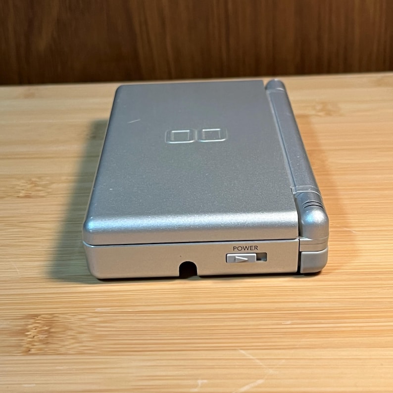 May include: A silver handheld gaming console in a closed position. The top cover features an embossed DS logo. The side edge displays a sliding power switch labeled with the word POWER and a small triangular icon indicating the direction to slide.