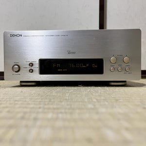 Puede incluir: Un sintonizador estéreo AM/FM Denon plateado, modelo UTUK-775, con una pantalla digital que muestra la frecuencia de radio FM. Tiene botones para encendido, reloj/pantalla, sintonización y funciones de memoria. El sintonizador está sobre una base plateada.