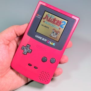 Puede incluir: Una consola de videojuegos portátil Game Boy Color de color rosa baya con una cruceta gris oscuro, botones A y B, y botones Select y Start. La pantalla muestra el título Hamtaro 2 en japonés con un personaje de hámster de dibujos.