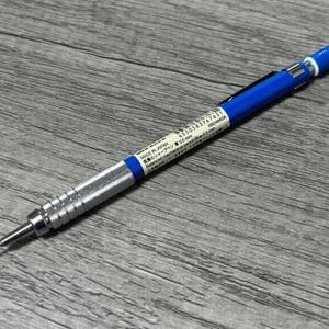 May include: A blue and silver mechanical pencil with a fine point. The pencil has a textured silver grip and a blue barrel. The pencil has a white label with Japanese text. The pencil is lying on a wooden surface.