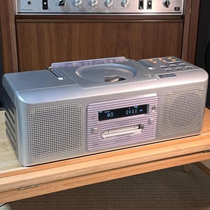 Könnte beinhalten: Ein silbernes tragbares Kenwood-Stereosystem mit einem CD-Player an der Oberseite, einem zentralen Digitaldisplay mit Anzeige von Titel 03 und Zeit 2:11, einem Kassettendeck und zwei seitlichen Lautsprechern mit perforiertem Metallgitter.