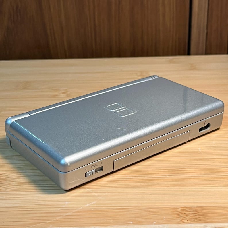 May include: A silver handheld gaming console with a rectangular, foldable design. The top cover features a debossed DS logo. The front edge includes a sliding volume control switch labeled VOL and a small headphone jack port on the side panel.