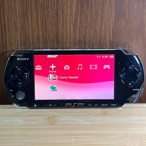 Puede incluir: Consola de juegos portátil Sony PSP negra con una cubierta protectora transparente. La pantalla muestra una interfaz rosa y roja con iconos para extras, cámara, música y vídeo. La consola tiene un pad direccional, botones y un joystick.
