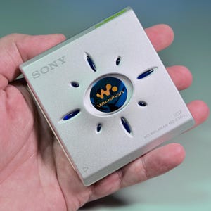 Puede incluir: Un reproductor de música portátil Sony MD Walkman MZ-E500 plateado de forma cuadrada. El dispositivo presenta un emblema circular azul central con el logotipo naranja de Walkman, rodeado por ocho pequeños acentos ovalados azules y el texto Digital SOUND PRESET.
