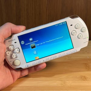 Może przedstawiać: Biała przenośna konsola do gier Sony PSP z jasnym ekranem wyświetlającym interfejs PlayStation Network. Urządzenie posiada pad kierunkowy, cztery przyciski akcji, okrągły analogowy drążek oraz różne przyciski sterujące.