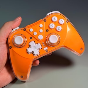 May include: A wireless video game controller featuring a bright orange transparent plastic shell. The device includes white analogue sticks, a white directional pad, and white action buttons labelled with X, Y, A, and B. Additional white function buttons are centred.