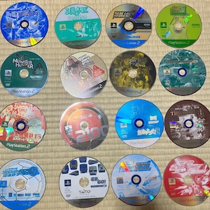 Puede incluir: Una colección de discos de videojuegos de PlayStation 2, cada uno con ilustraciones y títulos únicos. Los discos presentan una variedad de colores, incluyendo azul, verde, rojo y dorado. Algunos discos tienen texto en japonés. Los juegos se muestran sobre una estera tejida.
