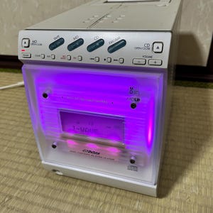 Puede incluir: Un equipo de música compacto plateado y blanco con un panel frontal iluminado en púrpura. El sistema tiene botones para cinta, MD, CD y FM/AM. La pantalla muestra "J-WAVE".