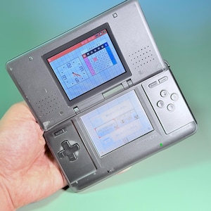 Peut inclure: Une console de jeu portable Nintendo DS argentée avec un design à double écran. L'écran supérieur affiche un calendrier pour septembre 2004 et une horloge analogique, tandis que l'écran tactile inférieur montre l'interface PictoChat.
