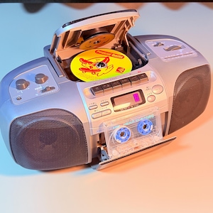 Può includere: Un lettore CD e cassette portatile color argento. Il lettore CD superiore è aperto e contiene un disco giallo con grafiche rosse. Una cassetta pulisci-testine trasparente è inserita nello scomparto frontale. Altoparlanti neri.
