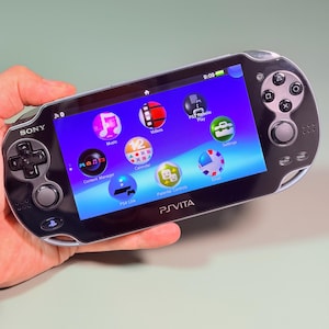 Könnte beinhalten: Eine schwarze Sony PlayStation Vita Handheld-Spielkonsole mit einem hellen Bildschirm, der ein Menü mit kreisförmigen Symbolen anzeigt, darunter Musik, Videos, PS3 Remote Play, Einstellungen, Inhaltsmanager, Kalender, E-Mail, PS4 Link und Kindersicherung.