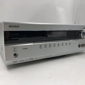 Op de afbeelding: Zilveren Onkyo-ontvanger met een zwart display en talrijke knoppen. Het voorpaneel heeft een koptelefoonaansluiting en een HDMI-ingang. Het Onkyo-logo is linksboven zichtbaar. Een grote zilveren knop bevindt zich aan de rechterkant.