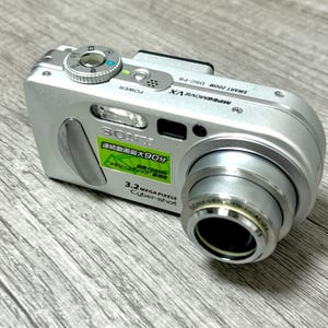 May include: Silver Sony Cyber-shot digital camera with a retractable lens. The camera has a flash, power button, and a dial on top. The lens is labeled "SONY LENS OPTICAL 3X". The camera has a green sticker with Japanese text.