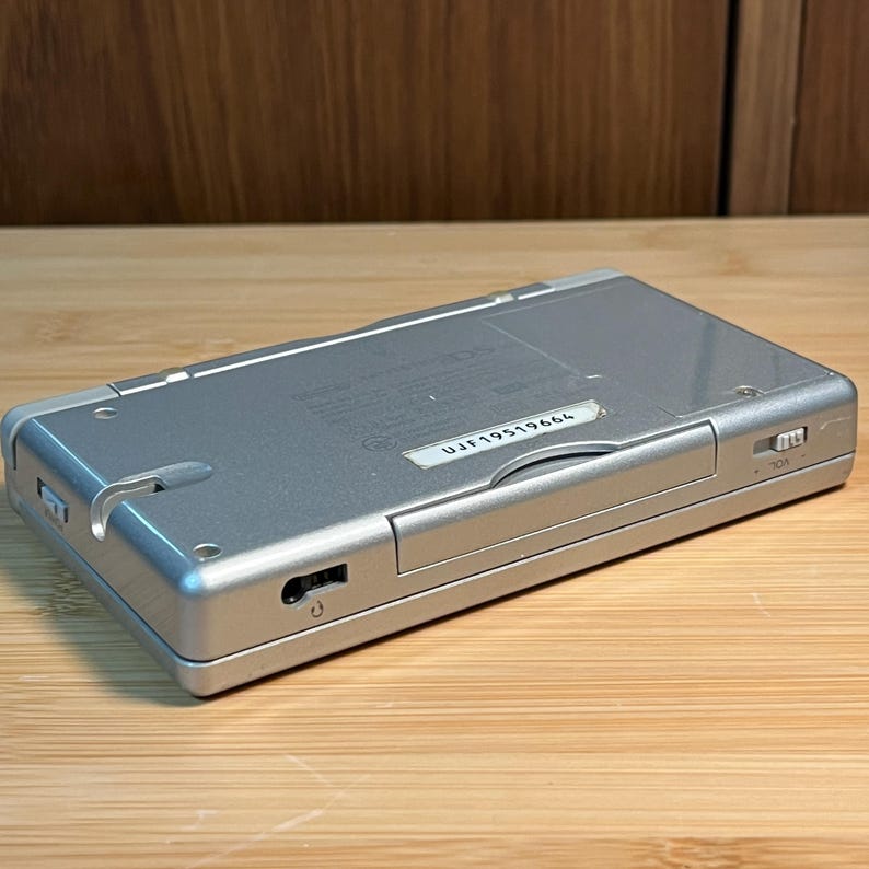 May include: A silver handheld gaming console featuring a rectangular body with a battery compartment, a volume slider labeled VOL, and a serial number sticker reading UJF19519664. The device includes a power switch and a headphone jack port.